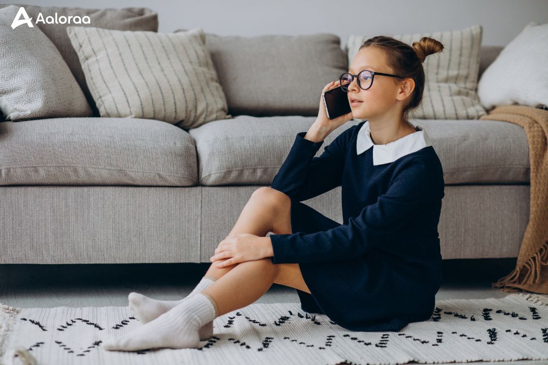 A young girl with glasses wearing a dark dress and gey socks sitting on a rug and talking on a smartphone in a living room
